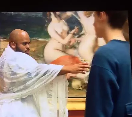 a black man in a white robe puts an arm out in front of a painting in a performance