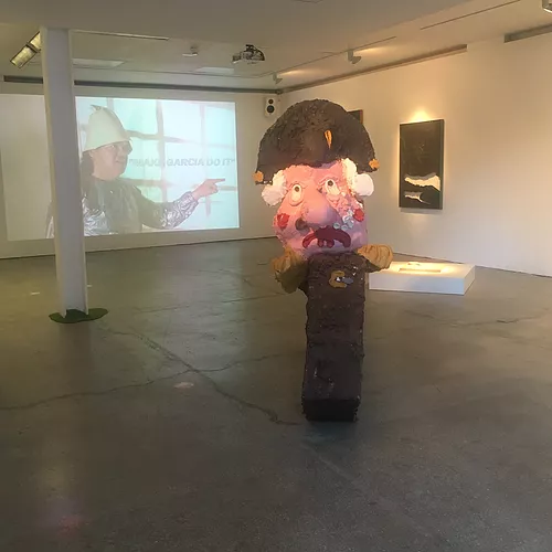 a papier mache looking face of an old man is the main sculpture in the centre of the space, and there&rsquo;s a dark painting on the wall in the back, and a projection of a person speaking too