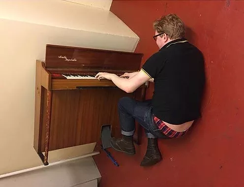 GT lies on the floor playing a piano that is up on its side on the wall