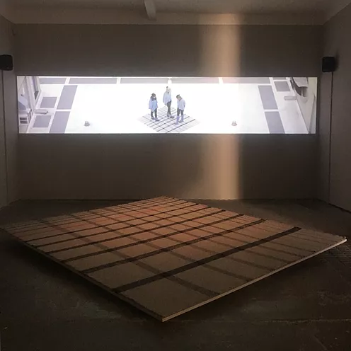 a very wide projection shows three people stood on a big square with a tartan pattern, and the same tarten square is on the floor of the gallery in front of the video 