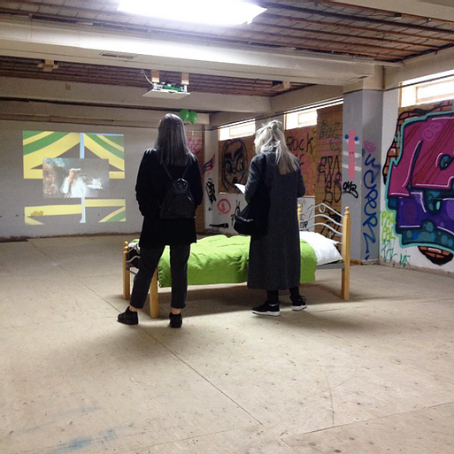 Gabrielle and Zarina stand watching a projection in a room with graffiti on the walls and a lone single bed in the middle of the space