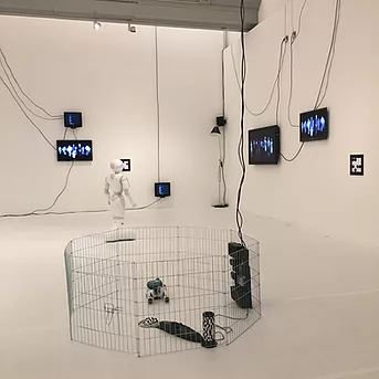another shot of the exhibition shows even more flat screens with wires flying across the walls, and a little robot dog on the floor in a metal pen
