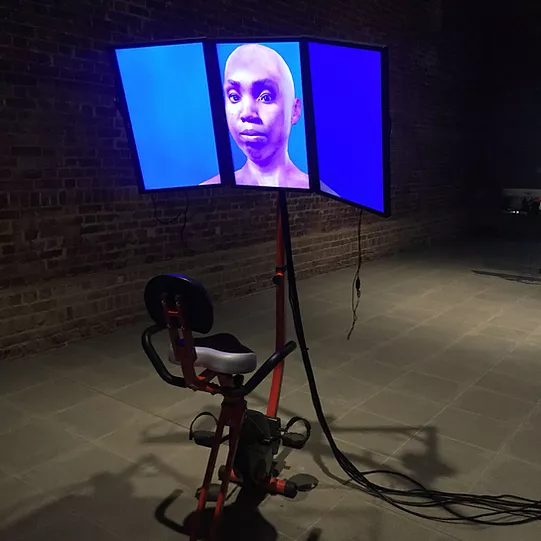 an exercise bike is place in front of three verticle screens placed next to each other so it looks curved, and there&rsquo;s a black woman against flat blue, and she has a blank expression