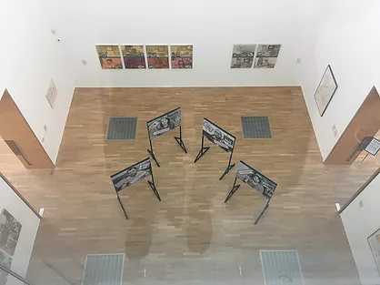 an image taken from the window upstais showing the gallery where different images are hung on walls, and there&rsquo;s also images on stands in the centre