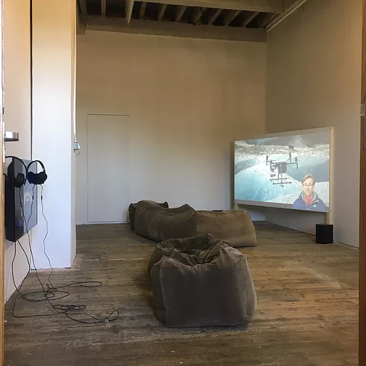 bean bags around a gallery space where there&rsquo;s a tv with headphones balancing on top of it, and a projection in the opposite corner as well