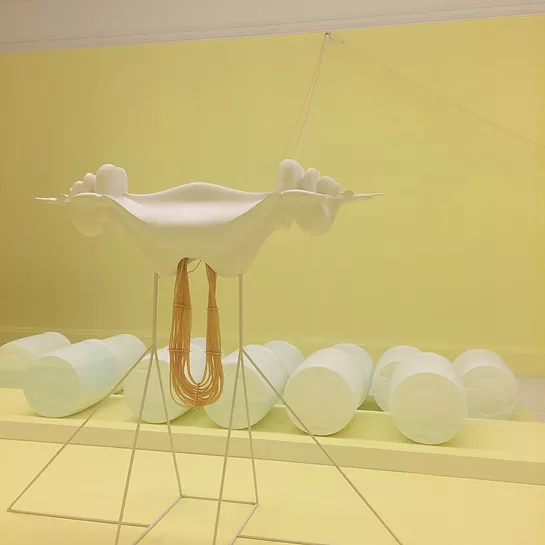 a strange bone-like sculpture with tubes hanging down below is propped up on meal frames with white cylinders in the back of a pale yellow gallery
