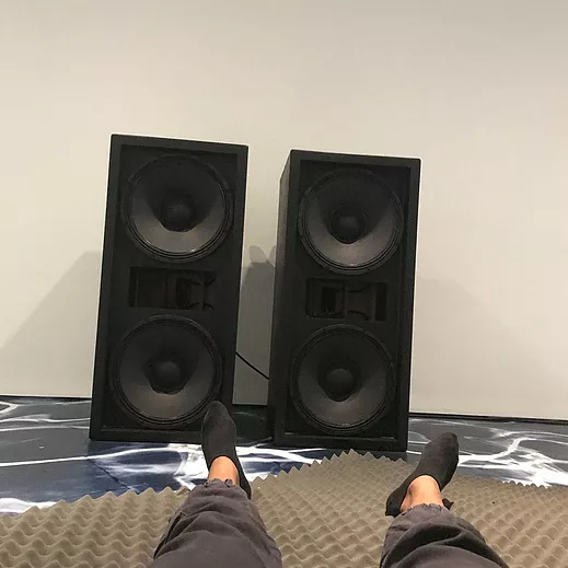 zarina lies on a gallery floor and takes a picture down at her feet which are visible in front of two big black speakers