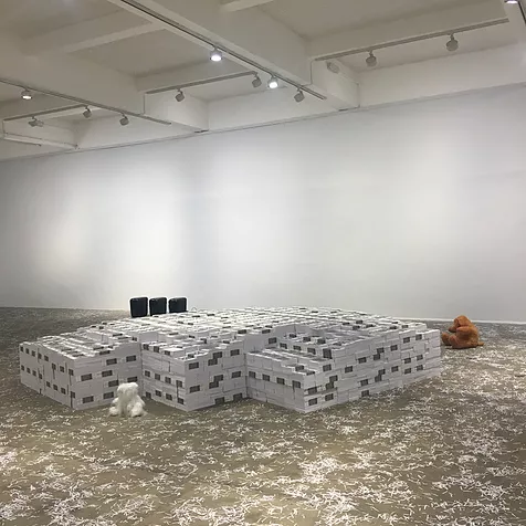 a huge white cube gallery space has massive piles of books in the centre of the space, some teddy bears at the side, and then the floor is littered with shredded paper from the books in the centre of the room