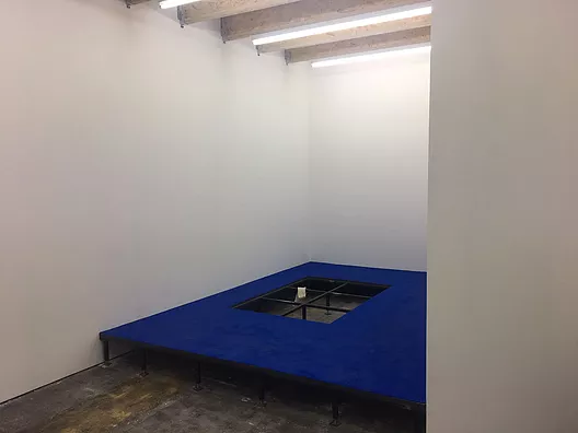 a white cube gallery space with a raised blue floor that has a gap in it, and there&rsquo;s a sock hanging over one of the bars revealed by the gap