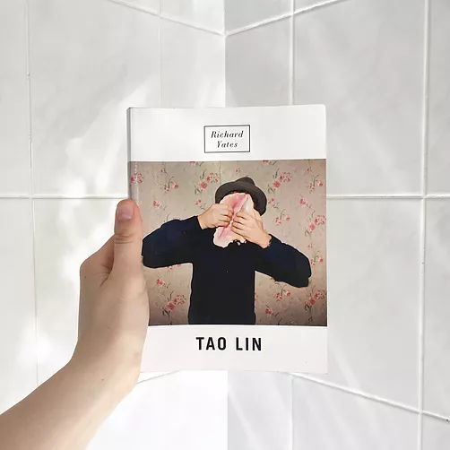 Gabrielle holds up the book Richard Yates by Tao Lin in the corner of a tiled bathroom. The front of the book has a man in a hat holding a giant shell in front of his face as if he&rsquo;s about to break it in half