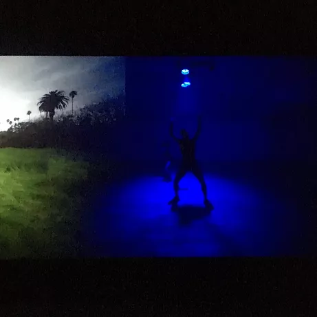the dual video shows palm trees on the left side, and a person performing in a room full of blue light on the right hand side