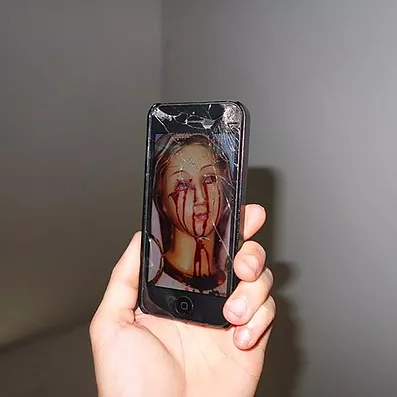 a white hand holds up a cracked iphone with an old painting of a woman with bright red blood pouring out of her eyes