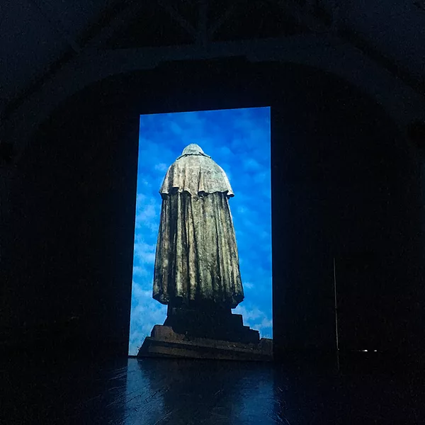 a huge portait projection shows a bright blue sky and a statue of a man with a robe on from behind on a plinth