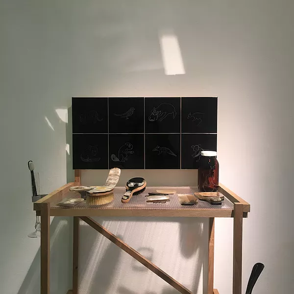a tiny makeshift table with a hair brush and a pot and a toothbrush on it, with black tiles where the mirror might be on a bedstand