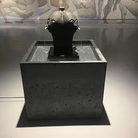 a low square water fountain with two black heads spurting water from pouting mouths, heads are conjoined at the back