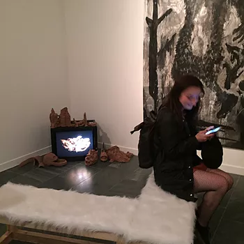 Gabrielle sits on the edge of a furry bench, and behind her there is a CRT in the corner with brown clay sculptures around it