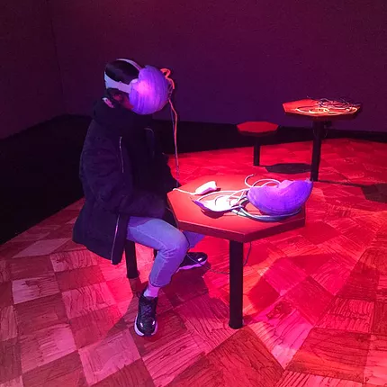 zarina sits at a tiny table with a big VR headset on that has been modified to look like a full purple round egg shape