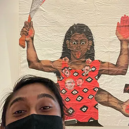 zarina takes a selfie with an artwork. the artwork looks crinkly, like it is an image plastered onto a wall. It depicts a brown woman with wide eyes sticking her tongue out, and she has four arms. we can see that the palms of her hands are painted red, and in one hand at the top she is holding a sword