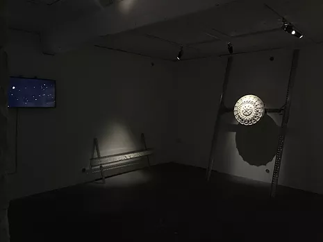 an installation shot showing a tv screen, a low plaster horizontal sculpture, and the ceiling rose which is held up at eye level with a wide metal structure leaning against the wall