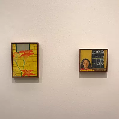 two small framed paintings on a wall alongside each other, one is orange lilies, and the other is a girl next to a window