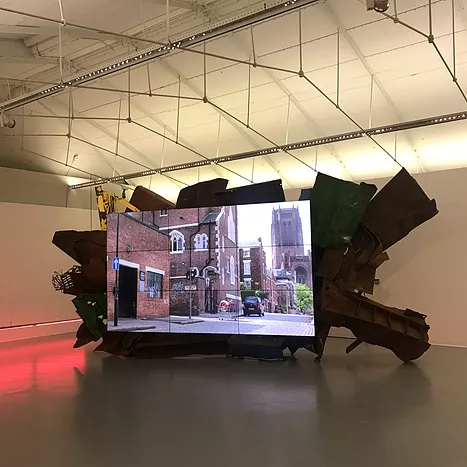 there is a huge screen showing a road leading up to the anglican cathedral in liverpool, and behind the screen there is an assemblage of scrap metal