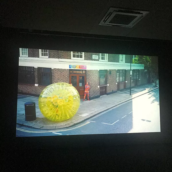 a big plastic yellow rubber sculpture is filmed on the corner of a road, and there is someone in an orange outfit just behind it at the entrance to the building, arms folded