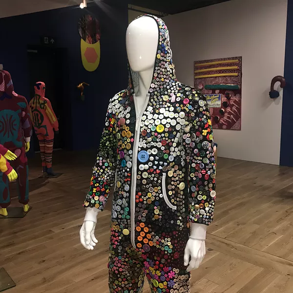a black tracksuit on a white mannequin is completely covered in random buttons stitched closely together, round ones, flowers, all different colours and shapes