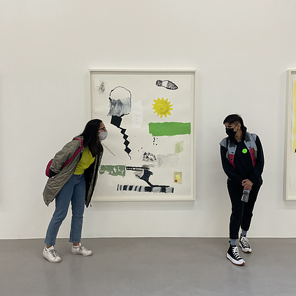 Amrita and Seema, two friends, pose either side of a white framed picture with small colourful details like a yellow sun, a green rectangle, a black stair case diagonal line and a shoe print