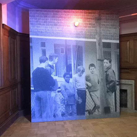 a black and white photo printed huge and leaning against a wood panelled gallery room - the image shows a bunch of kids leaning against a railing, one black kid amongst white kid, all boys, and on top of the image there is a real light, a red light fixed on top of them
