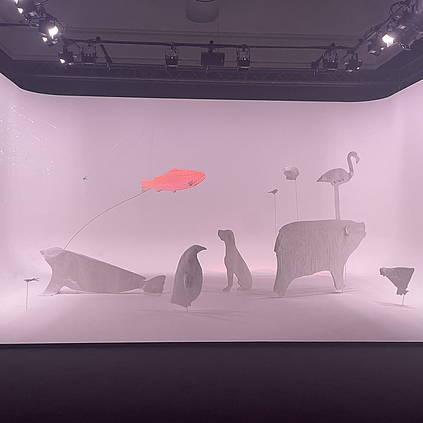 a big scene shows sculptures of animals in white grey shades - a flamingo, a dog, a penguin, a chicken, a seal - and then above them there is a salmon in pink flying over with a wire behind it holding it up in an arc