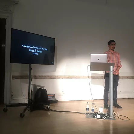 a man stands at a lecturn and a macbook pro, beside a TV screen that has the beginning of a presentation on it titled A Weight, A Course, A Coursing, Blood, A Ballast