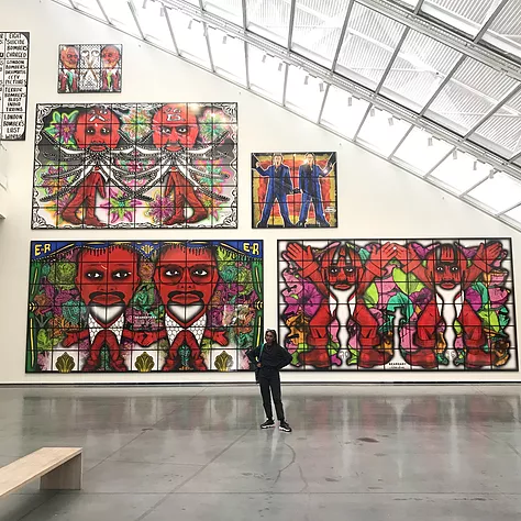 zarina stands with her hands on her hips in a huge white cube gallery space with reflective floors and a big slanted wall and ceiling. The wall is full of massive artworks by gilbert and george. The ones in this picture have repeated symmetrical bright red men on them