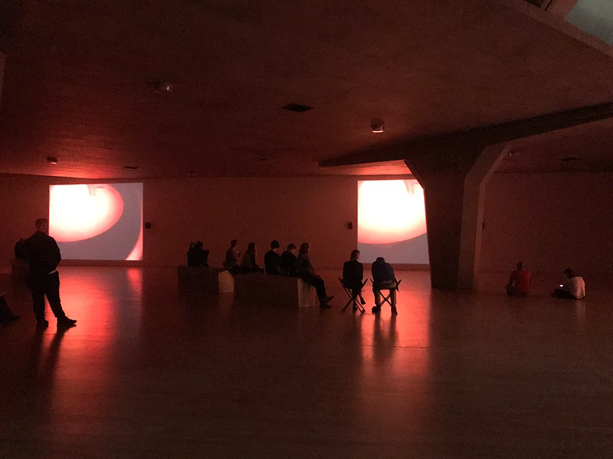 lots of people are sat around the gallery space on a bench, chairs, or on the floor, as the space is filled with an orange glow from projectors