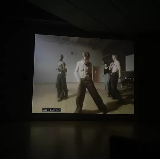 a topless white man with loose pants dances at the centre of the screen with people behind him