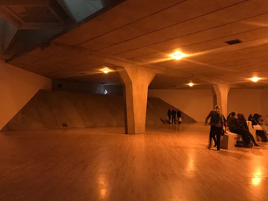 a room in tate with still visible wooden floors has been transformed so that it looks like the underpass of a motorway with columns, a concrete-like ceiling, and a huge sloping corner