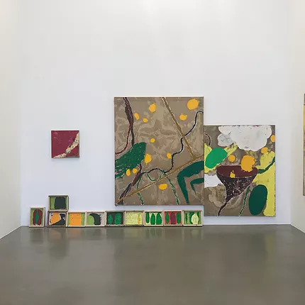 a collection of paintings low on a wall, side to side, and then multiple in a row right there on the floor. The paintings are earth tones, rough, scraped, and there are thin wavy lines against splodges and dots and an almost-cloud shape