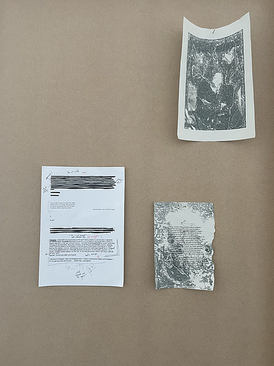 three pieces of paper are pinned on a wall; one is text with redactions, another is a photocopy of a book, and the other looks like a print but it is hard to tell what it is a print of