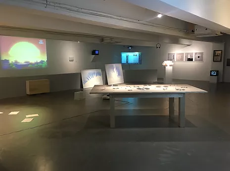a wide gallery space with low ceiling, there&rsquo;s a table in the centre with objects on it, and a long low gallery wall with paintings propped against it