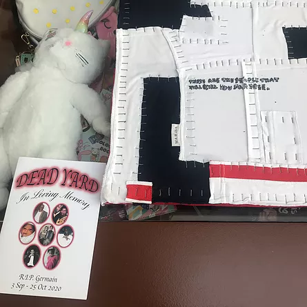 a stuffed animal that looks somewhere between a unicorn and a cat lies down next to a board that has white and black and red material stapled all along its edges in a close pattern, and there&rsquo;s a smaller A5 zine or piece of paper that says DEAD YARD and in loving memory of Germain with different images in circles on the front