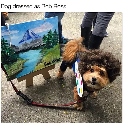 a dog is dressed like bob ross and next to him there is a tiny painting of a mountain and a forest on a tiny canvas. well done to the dog on his art