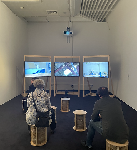 two people sit and watch an artwork that is split across three tv screens on wooden structures, all sitting on small wooden stools