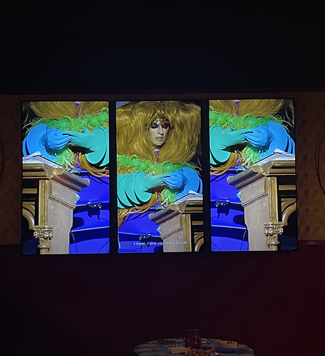 a video split into three portrait screens shows a drag queen with big wide yellow hair and rubber gloves on, the gloves are teal and blue respectively, and they stretch back in big concentric disc shapes
