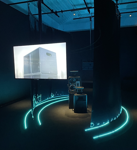 a dark gallery spaces shows a screen coming down from a wal with a big metal box on it, and it is situated in a big circular section with blue strip lights along the bottom
