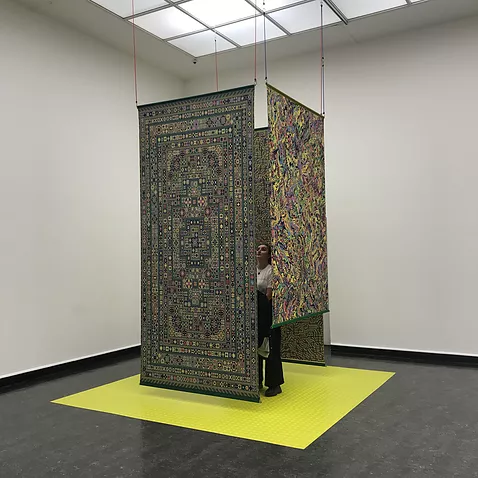 there is a cube of digitally woven carpets hung from the ceiling with a yellow square below and gabrielle is just about visible in the centre of them looking up at the details