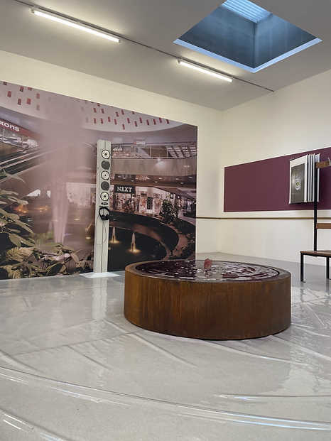 an exhibition shot shows a big image of a shopping centre with a Next store visible and a central fountain big on one wall, a circular low wooden fountain in the middle of the space, and the floor across the space has wrinkles, it&rsquo;s reflective, covered in some kind of thin plastic layer