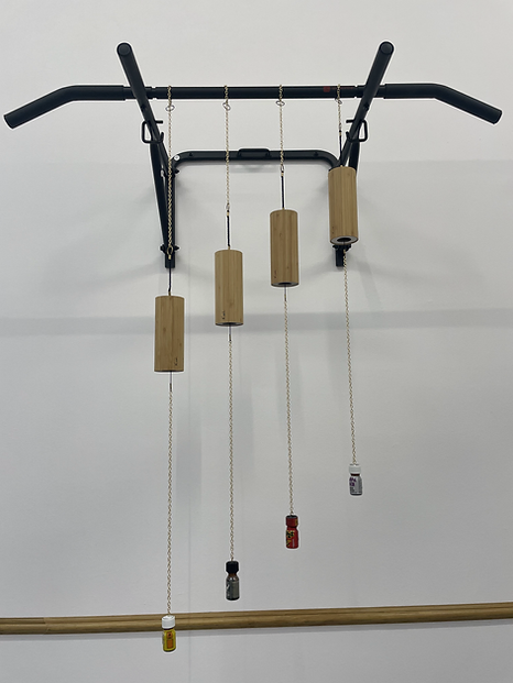 a metal frame high on a wall, like you might use to do pull ups, has four thin chains hanging down. half way down each chain there is a wooden cylinder, and at the very bottom a tiny charm of what appears to be stimulants
