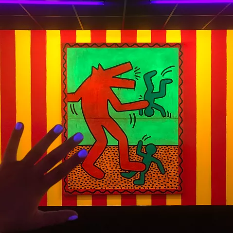 gabrielle holds her hand up to show white nailvarnish picked up by the UV light, in front of a painting of a standing red figure with a mouth like a dog, and two small green people, and the painting is hung on a red and yellow stripy wall
