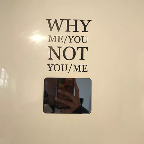 zarina takes a mirror selfie against a small bevelled edge mirror on the wall that has vinyl text above that says WHY ME/YOU NOT YOU/ME