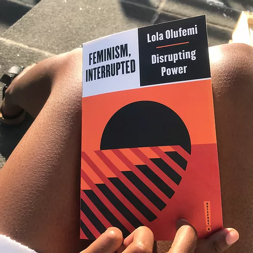 zarina is in a sunny place and showing the book she has on her lap which is Feminism Interrupted by Lola Olufemi, with a red black and orange graphic on the front like a black sunset disappearing into black bars