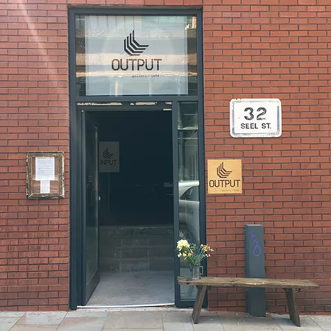 the output gallery facade shows a red brick wall, big frame door with a glass section at the top that says output with a spotlight behind it, and below there&rsquo;s a bench outside with a pot of flowers in it. All at 32 Seel Street